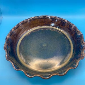 May include: A round ceramic pie dish featuring a scalloped edge. The interior surface displays a gradient of deep blue and earthy brown tones, while the outer rim is finished in a rich, glossy brown glaze with subtle variations in color.