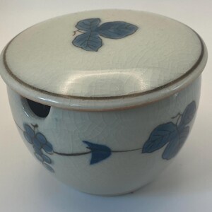 May include: A round ceramic lidded jar with a crackle glaze finish in off-white. The exterior features hand-painted blue grape clusters and leaves. The lid has a matching blue leaf motif, and the jar includes a small side notch for a spoon.
