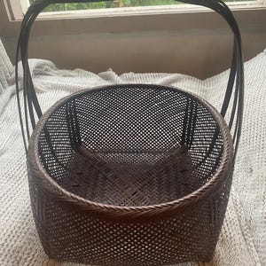 May include: A dark brown woven basket with a square base that transitions into a circular rim. The basket features a fine, intricate lattice pattern throughout its body and includes two tall, arched handles that extend upward.