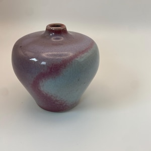 May include: A small, rounded ceramic vase with a narrow neck and a smooth, glossy finish. The glaze features a blend of muted mauve, dusty rose, and soft grey tones that swirl together across the surface of the vessel in a fluid, organic pattern.