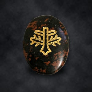 Puede incluir: Una piedra de obsidiana caoba de forma ovalada con una superficie pulida que presenta un patr&oacute;n moteado en marr&oacute;n oscuro y negro. Un s&iacute;mbolo grabado de color dorado que se asemeja a un &aacute;rbol estilizado o una cruz con halo est&aacute; centrado.