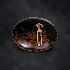 Puede incluir: Una piedra pulida de forma ovalada con una superficie moteada en negro y marr&oacute;n oscuro. En la parte frontal hay grabado un dibujo lineal de color dorado de una espada o daga estilizada, con una empu&ntilde;adura distintiva y una hoja larga y recta.