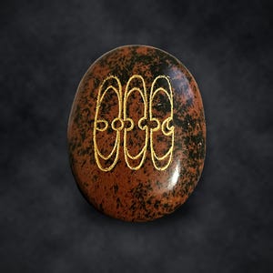 Puede incluir: Una piedra de obsidiana caoba de forma ovalada con una superficie lisa y pulida que presenta un patr&oacute;n moteado en negro y marr&oacute;n. El centro est&aacute; grabado con tres s&iacute;mbolos ovalados dorados interconectados en fila.