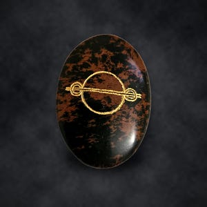 Puede incluir: Un cabuj&oacute;n de obsidiana caoba de forma ovalada con una superficie pulida de color marr&oacute;n oscuro y negro moteado. Un s&iacute;mbolo met&aacute;lico de color dorado que consiste en una barra horizontal que cruza un c&iacute;rculo con extremos en bucle est&aacute; centrado.
