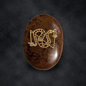 Puede incluir: Una piedra ovalada de color marr&oacute;n con una superficie pulida y moteada que presenta un dibujo lineal grabado en color dorado de una serpiente enroscada alrededor de un bast&oacute;n vertical. El dise&ntilde;o est&aacute; centrado en la piedra.