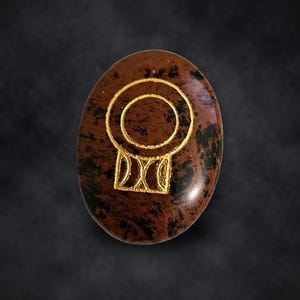 Puede incluir: Una piedra de obsidiana caoba de forma ovalada con una superficie pulida de color marr&oacute;n y negro moteado. Un s&iacute;mbolo grabado de color dorado est&aacute; centrado en la piedra, formado por un anillo circular sobre una base rectangular.