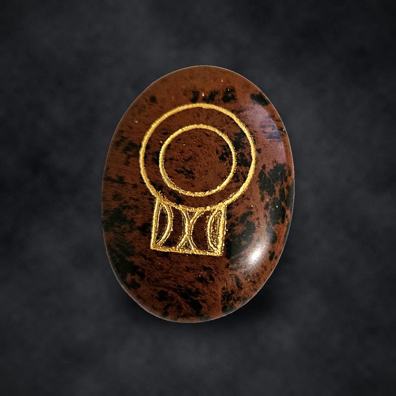 Puede incluir: Una piedra de obsidiana caoba de forma ovalada con una superficie pulida de color marr&oacute;n y negro moteado. Un s&iacute;mbolo grabado de color dorado est&aacute; centrado en la piedra, formado por un anillo circular sobre una base rectangular.