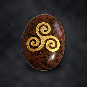 Puede incluir: Una piedra ovalada de color marr&oacute;n con una superficie pulida y moteada que presenta un s&iacute;mbolo de triskelion de color dorado grabado en el centro. La piedra muestra patrones jaspeados en marr&oacute;n oscuro y negro sobre su superficie lisa.