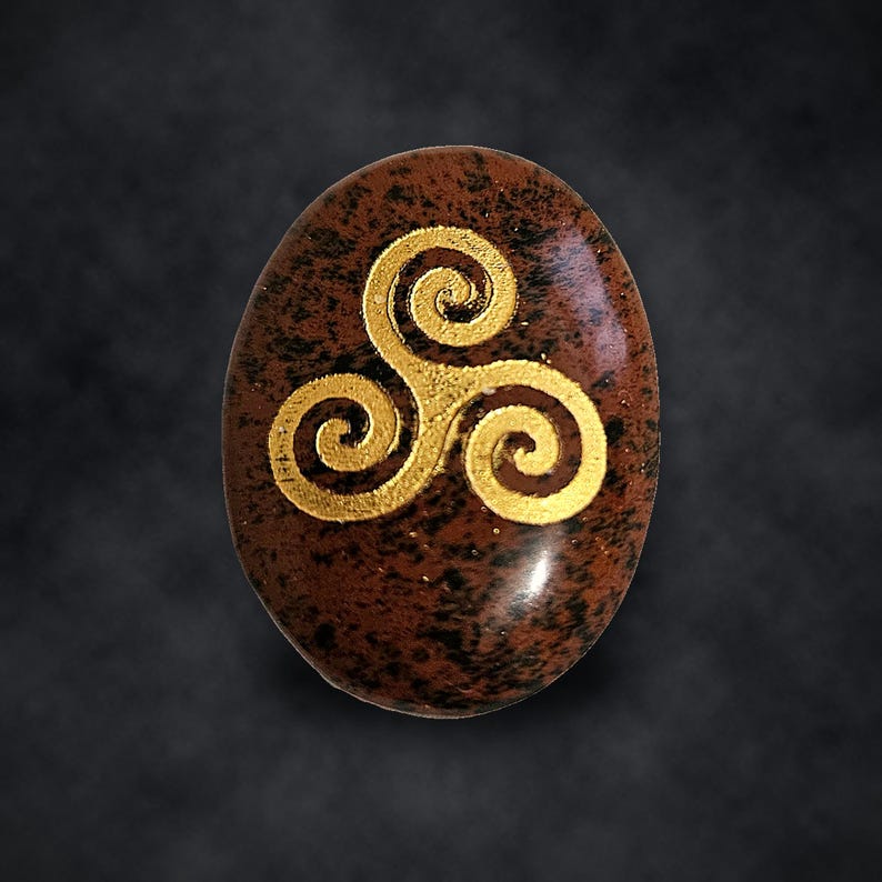 Puede incluir: Una piedra ovalada de color marr&oacute;n con una superficie pulida y moteada que presenta un s&iacute;mbolo de triskelion de color dorado grabado en el centro. La piedra muestra patrones jaspeados en marr&oacute;n oscuro y negro sobre su superficie lisa.