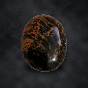 Puede incluir: Una piedra preciosa de obsidiana caoba pulida de forma ovalada, con una superficie lisa y brillante y un patr&oacute;n distintivo de remolinos de color negro profundo y marr&oacute;n rojizo que crean un aspecto jaspeado en toda la cara.