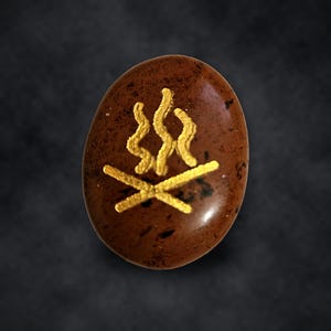 Puede incluir: Una piedra ovalada de color marr&oacute;n con un gr&aacute;fico dorado de una fogata. La piedra tiene una superficie lisa y pulida con patrones moteados en marr&oacute;n oscuro y negro, mientras que el dise&ntilde;o dorado representa dos troncos cruzados.