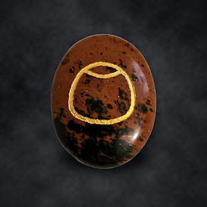 Puede incluir: Una piedra de obsidiana caoba de forma ovalada con una superficie pulida que presenta un patr&oacute;n moteado de color marr&oacute;n oscuro y negro. Un s&iacute;mbolo grabado de color dorado que se asemeja a un recipiente redondeado est&aacute; centrado.