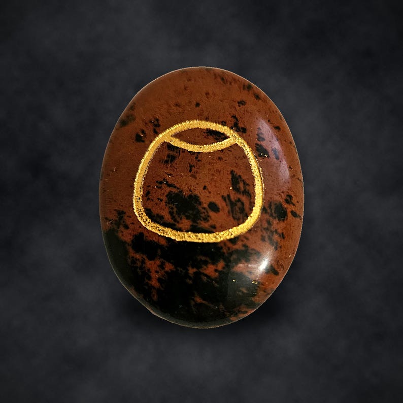 Puede incluir: Una piedra de obsidiana caoba de forma ovalada con una superficie pulida que presenta un patr&oacute;n moteado de color marr&oacute;n oscuro y negro. Un s&iacute;mbolo grabado de color dorado que se asemeja a un recipiente redondeado est&aacute; centrado.