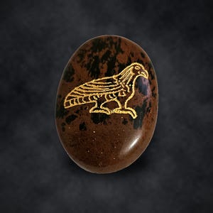 Puede incluir: Una piedra ovalada de color marr&oacute;n con patrones negros moteados que presenta un gr&aacute;fico grabado en color dorado de un ave rapaz de perfil. La piedra tiene una superficie lisa y pulida con el dise&ntilde;o del ave grabado en el centro.