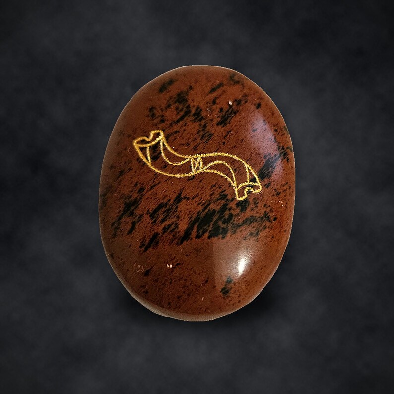 Puede incluir: Una piedra ovalada de color caoba con una superficie lisa y pulida que presenta patrones moteados en marr&oacute;n oscuro y negro. Un dibujo lineal de color dorado de un cuerno curvado est&aacute; grabado en el centro de la cara frontal de la piedra.