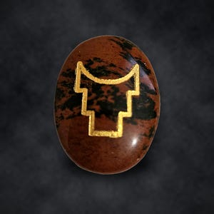 Puede incluir: Una piedra preciosa de obsidiana caoba de forma ovalada con una superficie pulida y lisa con patrones naturales de remolinos negros y marrones. Un s&iacute;mbolo geom&eacute;trico dorado en forma de U con lados escalonados est&aacute; grabado en el centro.