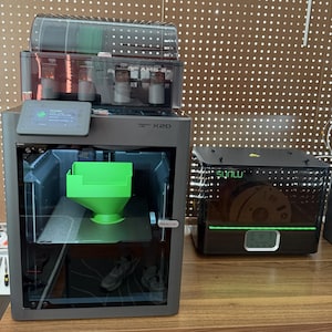 Może przedstawiać: Ciemnoszara drukarka 3D Bambu Lab X1C z jednostką AMS na górze, zawierająca jasnozielony obiekt w kształcie lejka na stole roboczym. Obok znajduje się czarny osuszacz filamentu Sunlu z zielonym paskiem LED i cyfrowym wyświetlaczem.