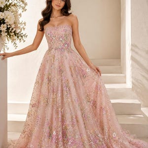 May include: A strapless floor-length gown in a soft pink hue, featuring a fitted bodice and a full, voluminous skirt. The entire garment is embellished with intricate floral embroidery in shades of pink, green, and gold, accented with sequins.