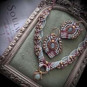 Op de afbeelding: Een macramé ketting en bijpassende oorbellenset met ingewikkelde lichtblauwe en mauve koordpatronen, kleine metallic gouden kralen en centrale lichtblauwe cabochonstenen. De ketting bevat een goudkleurige metalen hanger met een steen.