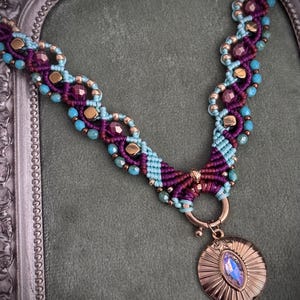 Op de afbeelding: Een macramé ketting van donkerpaars en lichtblauw koord, geweven met metallic gouden kralen en gefacetteerde paarse kralen. De ketting heeft een koperkleurige ronde hanger met een centrale iriserende steen in marquisevorm.