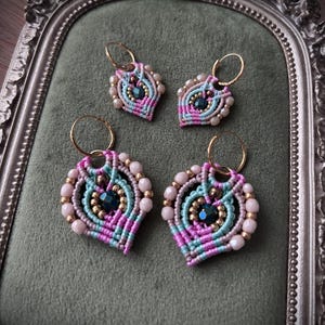 Op de afbeelding: Twee paar macramé oorbellen gemaakt van geknoopt koord in roze, lichtblauw en grijs. Elke oorbel is versierd met kleine ronde kralen in zachtroze en metallic goud, met een donkerblauwe gefacetteerde kraal in het midden aan een gouden ring.