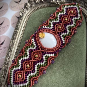 Op de afbeelding: Een macramé armband met een centrale witte ovale steen, ingewikkelde geweven koordpatronen in koper, paars, wit en olijfgroen, kleine goudkleurige kralen, paarse gefacetteerde accenten en een kleine ronde goudkleurige bedel met bloemmotief.