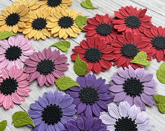 May include: Assortment of paper craft die cuts featuring various colors of sunflowers with black centers. The sunflowers are arranged in a circular pattern with green leaves scattered around them.