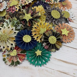 May include: A collection of paper craft flowers with glitter accents. The flowers are in various colors and patterns, including green, blue, yellow, and brown. The flowers are arranged in a pile on a white wooden surface.