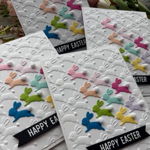 May include: Four white cards with a raised embossed design of bunnies with fluffy tails. The bunnies are in various colors, including pink, blue, green, and yellow. Each card has a black banner with the text "Happy Easter" printed on it.