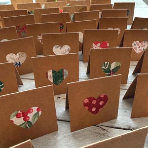 May include: A collection of folded brown paper cards, each featuring a heart-shaped cutout revealing a patterned design. The designs include floral, berry, and holiday-themed patterns. The cards are arranged on a light-colored wooden surface.