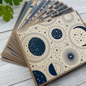 May include: A stack of blank greeting cards with a celestial pattern. The cards feature a cream background with navy blue moons, stars, and celestial bodies.
