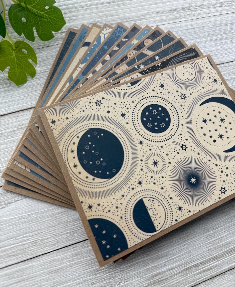 May include: A stack of blank greeting cards with a celestial pattern. The cards feature a cream background with navy blue moons, stars, and celestial bodies.