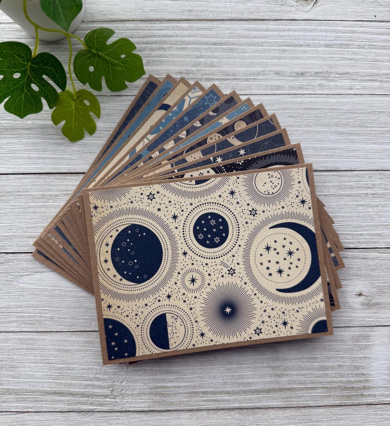 May include: A stack of blank note cards with a celestial design featuring moons, stars, and a navy blue and cream colour scheme.