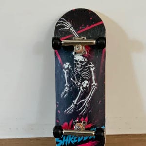 May include: A fingerboard featuring a black deck with a white skeleton graphic, pink and blue paint splatters, and the text SHREDDER in blue. The board includes silver metal trucks and black wheels, with a small logo in the top left corner.