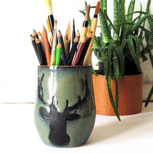 May include: A ceramic pencil holder with a deer silhouette design in black on a teal-green background. The holder is filled with various colored pencils and paintbrushes. A potted aloe vera plant is in the background.