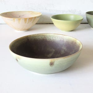 May include: A collection of ceramic bowls in various colors including a large pale green bowl with a dark brown interior, a light pink bowl with drip glaze, a bright lime green bowl, a dark olive green bowl, and a partial teal bowl.