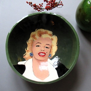 Puede incluir: Un tazón de cerámica verde con un retrato pintado de una mujer con cabello rubio y labios rojos. La mujer lleva pendientes azules.