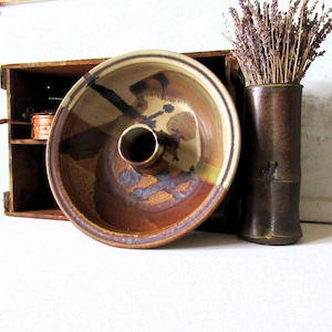 May include: A round ceramic bundt pan with a central tube, featuring a glazed finish in shades of brown, tan, and dark charcoal. Beside it stands a tall, cylindrical dark brown ceramic vase filled with a bundle of dried lavender stems.