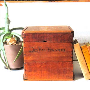 Puede incluir: Una caja rectangular de madera con tapa abatible, que presenta un acabado marrón cálido y clavos metálicos visibles a lo largo de los bordes. El panel frontal muestra el nombre John Havens escrito a mano con tinta negra sobre la madera.