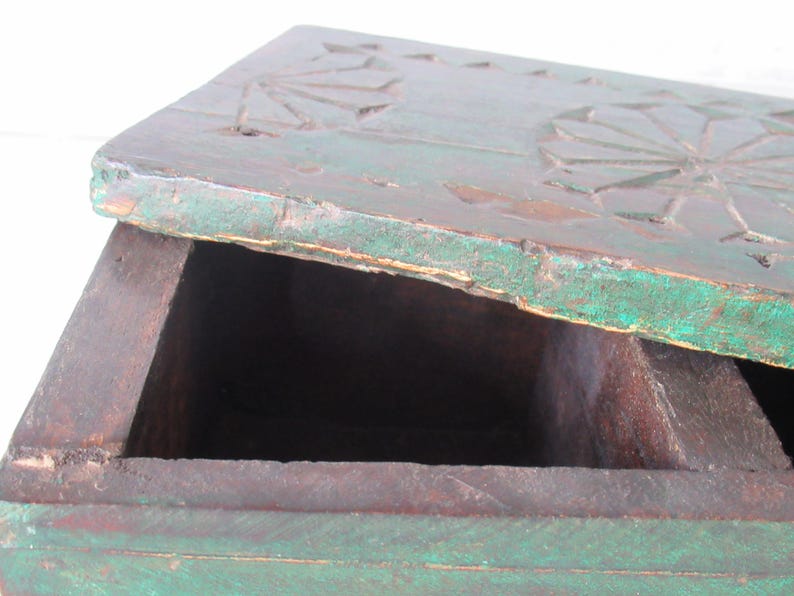Puede incluir: Caja de madera antigua parcialmente abierta, con acabado turquesa desgastado. La tapa presenta dise&ntilde;os florales tallados. El interior es oscuro, revelando compartimentos. Probablemente utilizada para almacenamiento o exhibici&oacute;n.