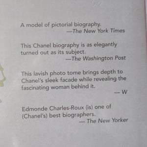 May include: Close-up of a book cover with text. The cover is light pink with text in black, including quotes from The New York Times, The Washington Post, and The New Yorker. The book is a biography about Chanel.
