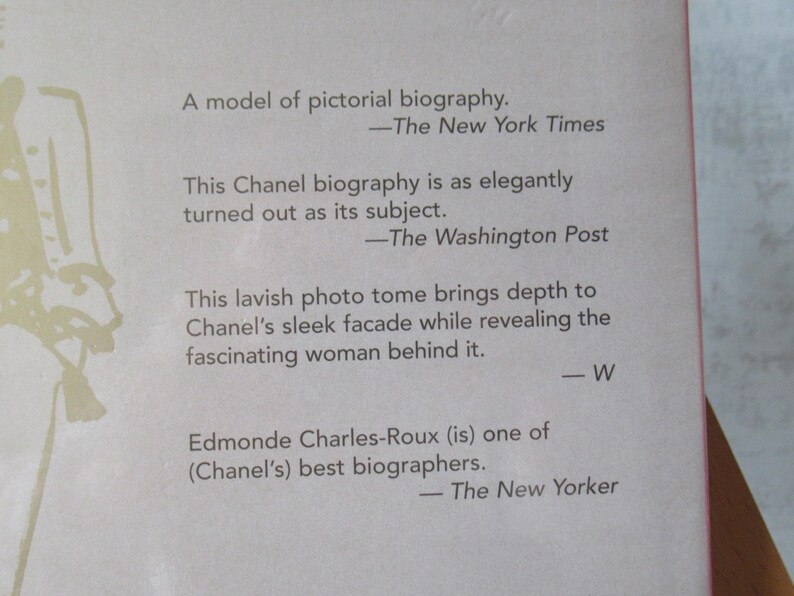 May include: Close-up of a book cover with text. The cover is light pink with text in black, including quotes from The New York Times, The Washington Post, and The New Yorker. The book is a biography about Chanel.