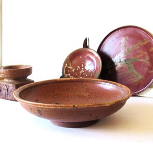May include: A collection of ceramic tableware including a wide, shallow, speckled reddish-brown bowl in the foreground, a smaller matching dish behind it, a large dark red plate with green accents, and a carved wooden pedestal base to the left.