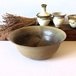 May include: A round, wide-rimmed ceramic bowl with a smooth, matte, earthy tan exterior and a darker, mottled interior glaze. The bowl is positioned in front of three smaller, cream-colored ceramic vessels and a bundle of dried lavender.