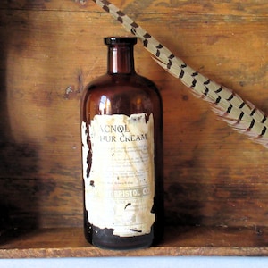 May include: A vintage brown glass bottle with a label that reads "ACNOL HUR CREAM" and "BRISTOL CO". A long feather with brown and black markings rests on a wooden surface.
