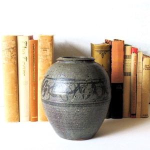 Könnte beinhalten: Eine graue Keramikvase mit einem dunkelblauen abstrakten Design in der Mitte. Die Vase steht vor einer Sammlung von Vintage-Büchern mit unterschiedlich farbigen Buchrücken, darunter Beige, Orange und Gelb.