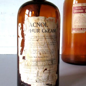 May include: Two vintage amber glass bottles with aged labels. One bottle reads "ACNOL Sulphur Cream" and the other "Cascara Evacuant" from Parke, Davis & Co. The labels show signs of wear and tear, adding to their antique charm.