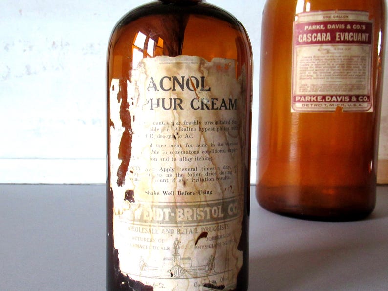 May include: Two vintage amber glass bottles with aged labels. One bottle reads "ACNOL Sulphur Cream" and the other "Cascara Evacuant" from Parke, Davis & Co. The labels show signs of wear and tear, adding to their antique charm.