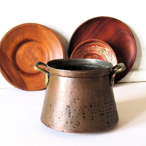 May include: A hammered copper pot with two brass side handles, positioned in front of three wooden plates. The pot has a wide, tapered body with a metallic finish and visible dark patina spots, while the plates are round and brown.