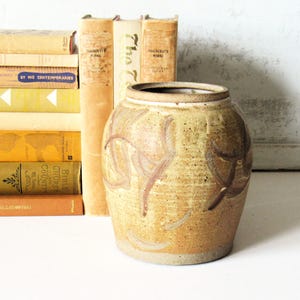 May include: A round, tan ceramic jar with a speckled texture and brown painted leaf-like motifs. The jar has a wide, open mouth with a thick, unglazed rim. It is positioned in front of a stack of several vintage books with yellow and brown spines.